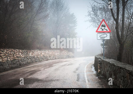 Slippery road in rainy weather, warning roadsign stands on mountain roadside, rural landscape Stock Photo