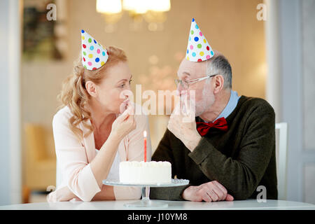 Mature man and woman in birthday caps tasting yummy dessert Stock Photo