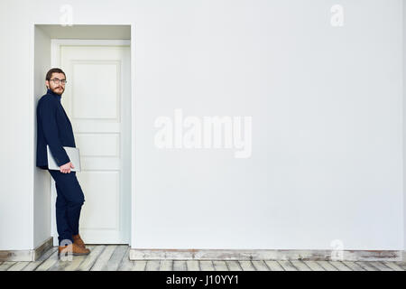 Bearded man in glasses leaning on doorway holding laptop computer and looking at camera next to blank white wall in office Stock Photo