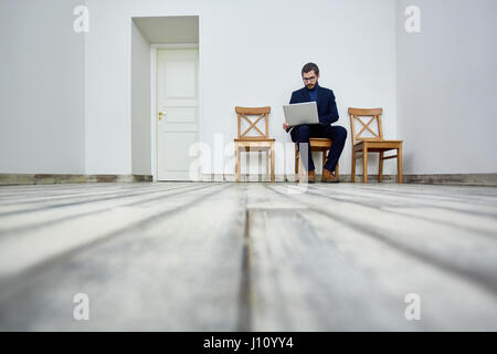 Low angle portrait of bearded man using laptop while sitting on chair in waiting room by office door Stock Photo