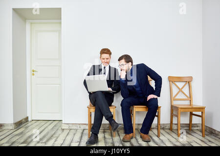 Portrait of two business people using laptop sitting on chairs in waiting room outside office Stock Photo