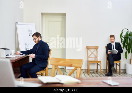 Two business people working in old-school business office, one man reading documentation at desk, other man using tablet  sitting on chair Stock Photo