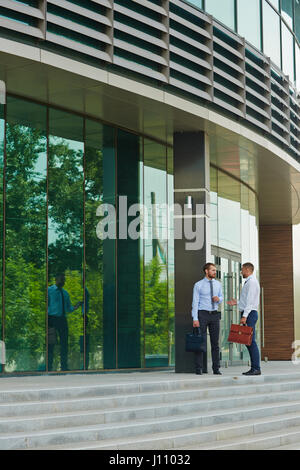 Two young businessmen talking on steps of modern office building with glass fronts Stock Photo