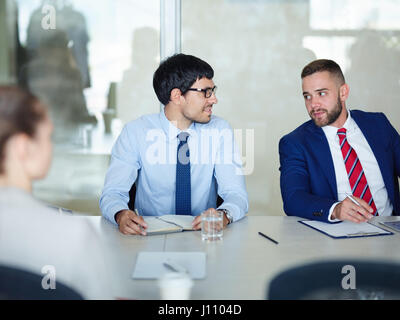 Portrait of two young business people leaning to each other while talking at meeting table, discussing corporate issues Stock Photo