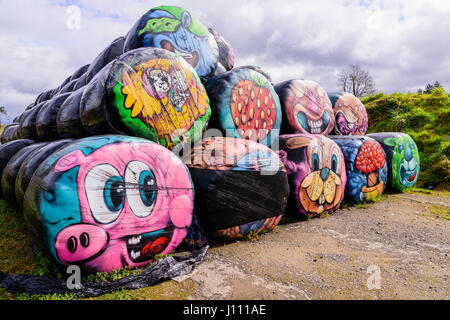 Haybales stacked at the side of a road with various colourful faces and objects painted on them. Stock Photo