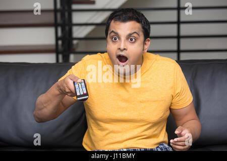 Closeup portrait, young man in yellow t-shirt, sitting on black leather couch, watching TV, holding remote, surprised at what he sees, mouth wide open Stock Photo
