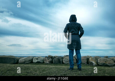 Lonely hooded female person from behind standing at seashore and looking into distance on a cold winter day, concept of waiting, anticipation, hope an Stock Photo