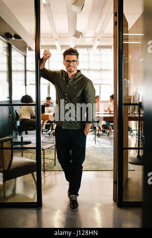 Full length portrait of happy young businessman standing in doorway of office. Caucasian male executive in office with people working in background. Stock Photo
