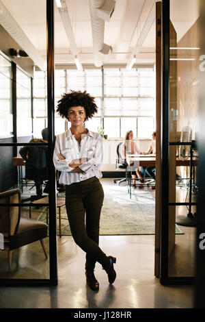 Full length portrait of confident young woman standing in doorway of office with her arms crossed. Creative female executive at startup with people wo Stock Photo