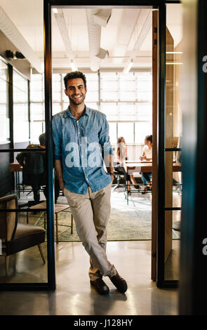 Full length portrait of handsome young man standing in doorway of office with his hands in pockets. Creative male executive at startup with colleagues Stock Photo