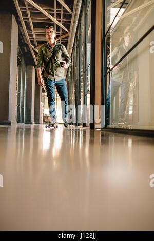 Full length shot of young man skateboarding in office. Casual businessman skating through office corridor. Stock Photo