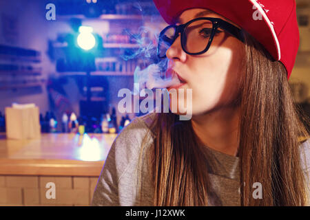 Young pretty woman in red cap smoke an electronic cigarette at the vape shop. Closeup. Stock Photo