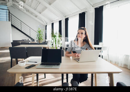 Young woman sits at the cafe table using a laptop and holding a cell phone while looking away with thoughtful gaze. Stock Photo