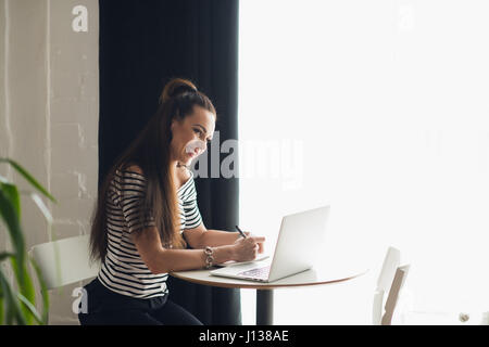 Photo of pretty woman making notes and thinking over a subject while sitting in some coffee shop with her laptop near big window. Stock Photo