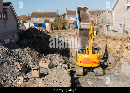 Excavator is digging a house foundation in a residential area while a dumper truck is unloading construction gravel, sand and crushed stones on the co Stock Photo