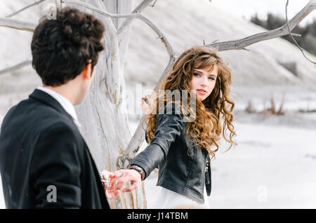 Young woman with curly hair holding hand of young man standing back to the camera close to the withered tree in the desert. Stock Photo
