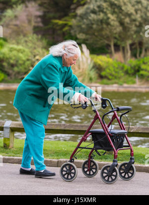 Elderly lady walking with a rollator (wheeled walker or wheeled Zimmer frame) by a lake in a park in England, UK. Stock Photo