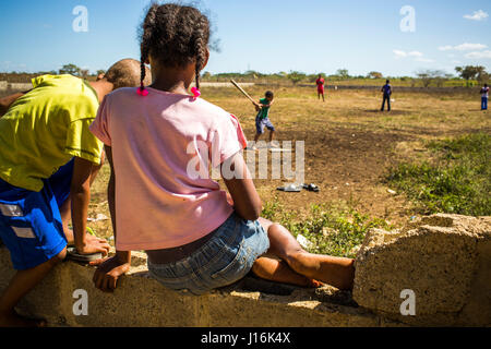 Girl With Ponytails Sits On A Wall Overlooking A Sandlot And People Playing Baseball On A Sunny Day Stock Photo