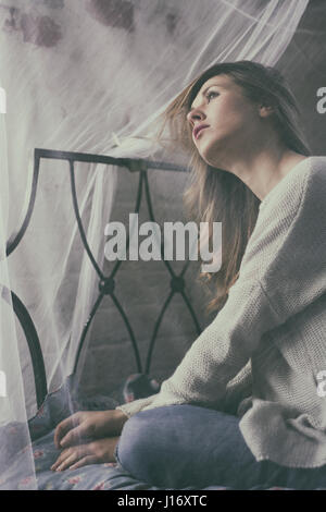 Pensive young woman sat on bed looking up Stock Photo