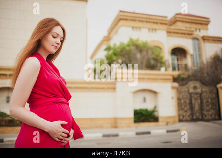 Cute red-haired pregnant girl in a red dress. On the background of the Villa. Stock Photo