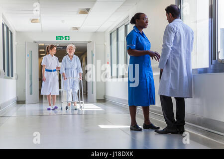 Nurse helping a female elderly old woman patient using walking frame in a hospital corridor with a doctor Stock Photo