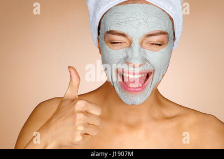 Charming young girl with mask of clay on her face. Photo of crazy girl shows thumbs up and smiling. Beauty & Skin care concept Stock Photo
