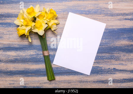 A bouquet of daffodils and a sheet of white paper on a wooden background. Spring yellow flowers. Paper with space for text. Blue peeling paint. Flat l Stock Photo