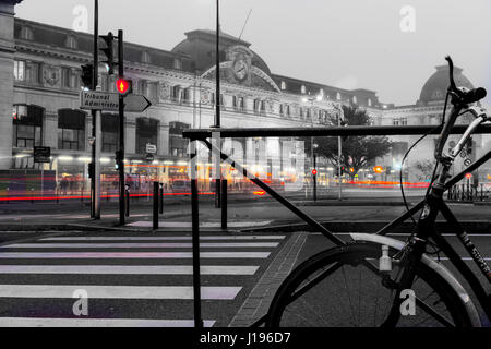 Cycle leaning against railing under morning mist in city with glow of lights on street, path and buildings with Matabiau Railway Station in background Stock Photo