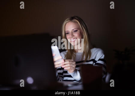 Young beautiful woman sitting at desk at night, laptop in front of her, holding smart phone, texting. Stock Photo
