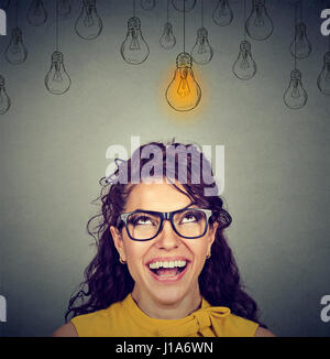 Portrait happy beautiful woman in glasses looking up at idea light bulb above head isolated on gray wall background Stock Photo