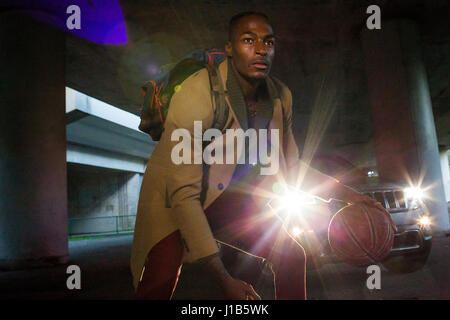 Headlights shining on Black man wearing backpack playing basketball Stock Photo