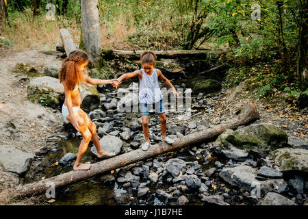 Mixed Race sisters holding hands on log over forest stream Stock Photo