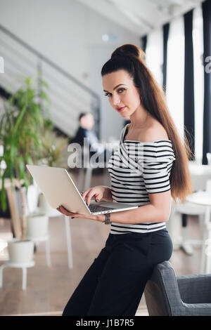 Beautiful adult female is holding a laptop and looking at the camera while standing in an empty cafe. Stock Photo
