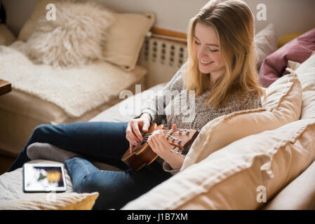 Smiling woman sitting on sofa playing ukulele Stock Photo