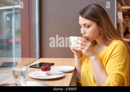 A young woman is taking a sip of coffee at the table with raspberry cake, glass of water and tablett on it. The scene is next to a window. Stock Photo