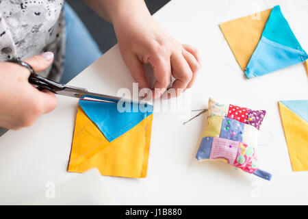 patchwork and quilting in the workshop of tailor woman - close up on tailor scissors cuts the thread on the blue and yellow scraps of fabric for patchwork on the desktop with thread, pins and cushion. Stock Photo