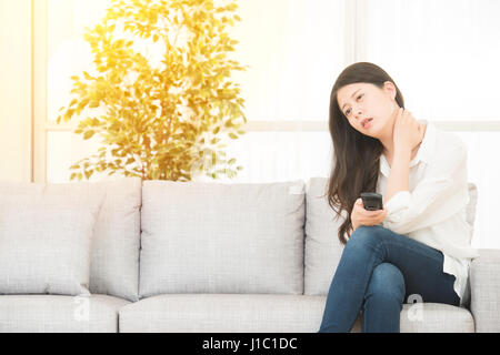 beautiful woman suffering from painful neck sitting on cosy sofa watching TV. lifestyle and health concept. mixed race asian chinese model. Stock Photo