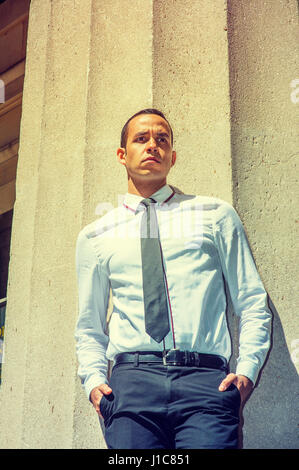 Young American Businessman wearing white shirt, black tie, standing against column outside office building in New York, under strong sun, frowned, thi Stock Photo