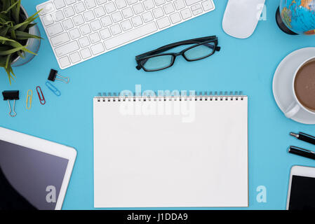 Modern blue office desk table with computer, tablet, cellphone, other supplies, eye glasses, cup of coffee and blank notebook page for input the text. Stock Photo