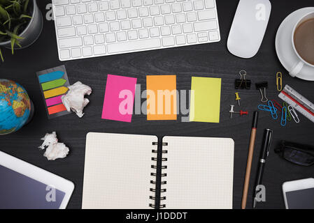 Office table desk with white blank note pad, supplies, coffee cup, pen, pencil, computer, crumpled paper, flower and eye glasses on wooden black backg Stock Photo
