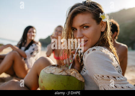 Portrait of attractive young woman drinking fresh coconut water with her friends sitting in background on the beach. Young people on summer vacation. Stock Photo