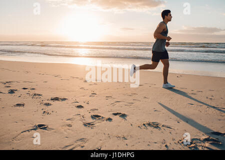 Side view shot of fit young man running on the beach in morning. Healthy male model jogging on the sea shore. Stock Photo