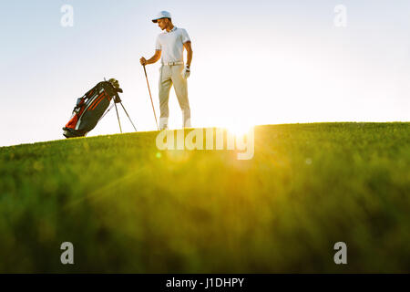 Full length of golf player looking at bag while standing on field during sunny day. Low angle shot of male golfer on golf course. Stock Photo