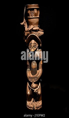 This image features a highly rare, masterfully carved 19th-century traditional Chokwe drum player figure or standard, created by an artisan from Angola or the borderlands of the Democratic Republic of the Congo and Zambia. Chokwe wooden standard depicting a standing figure balancing a ceremonial drum on their head, West Central African sculpture isolated on a black background Stock Photo