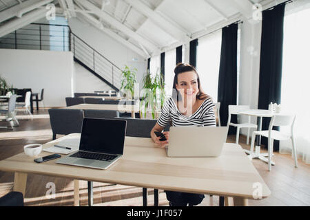 Portrait of an attractive woman sitting at the table with a laptop computer holding phone and looking away searching for inspiration for her book. Stock Photo