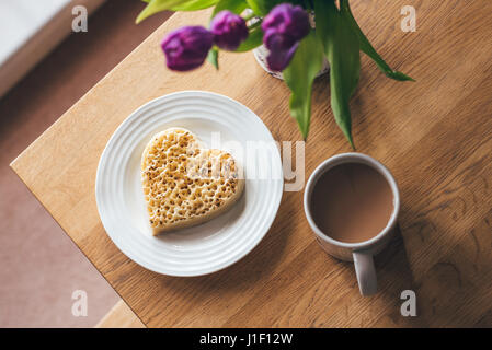 Heart-shaped toasted crumpet on white plate with mug of tea on table with purple tulips. Stock Photo
