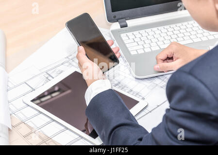 close up of businessman holding smartphone with black screen over tablet pc computer notebook blueprints paper and blueprint rolls on the table in the Stock Photo