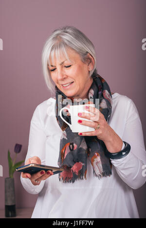 Smiling grey-haired woman using smartphone while drinking coffee. Stock Photo
