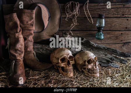 Still life with two human skulls on hay with traditional leather boots and american west rodeo brown felt cowboy hat background, vintage and dark tone Stock Photo