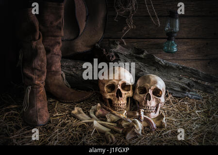 still life with human skull on hay and animal bones in traditional leather boots and american west rodeo brown felt cowboy hat background, vintage and Stock Photo